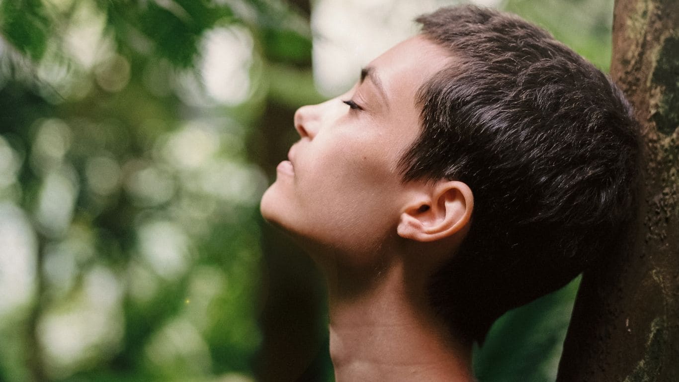 Imagem de uma mulher de cabelos curtos de perfil e de olhos fechados em meio à natureza