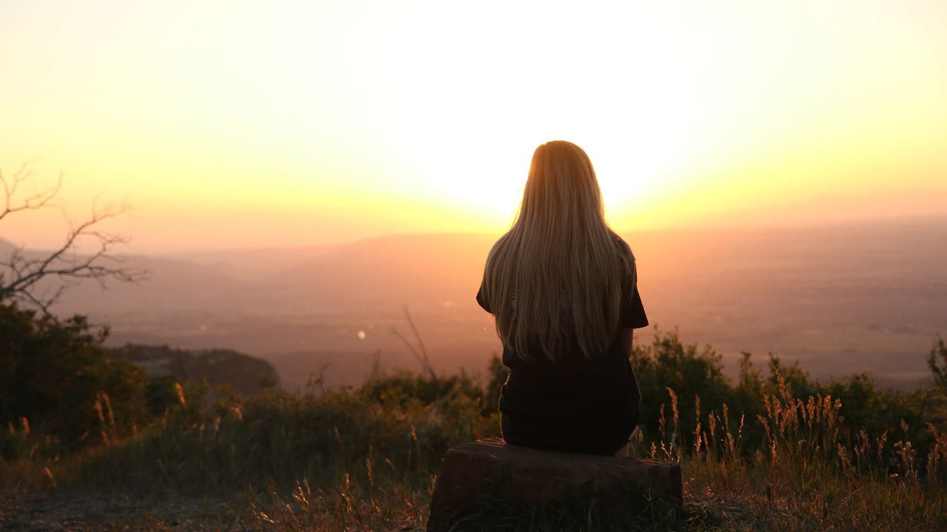 Imagem de uma mulher sentada de costas olhando o entardecer entre as montanhas